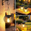 A close-up of a solar powered metal hanging lantern with a wire mesh design illuminating an outdoor setting.