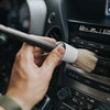 A hand is using a soft detailing brush to clean the interior of a car stereo or dashboard.