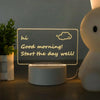 A white acrylic message board desk lamp displays a glowing text message "hi Good morning! Start the day well!" on a dark surf
