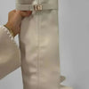 A close-up of a beige knee-high boot with a fabric strap and metal buckle being held by a hand.