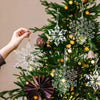 Hånd henger klar akryl-snøfnuggpynt på juletre med lys og grønt
