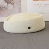 A cream-colored straw pillbox sun hat with a wide brim and a gold initial charm resting on a gray surface.