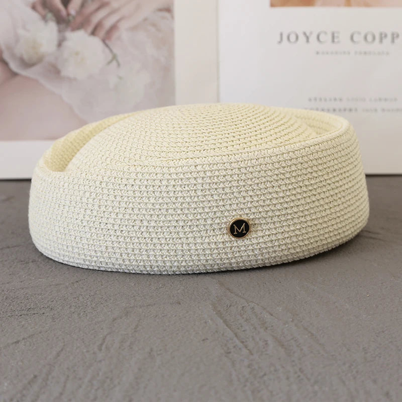 A cream-colored straw pillbox sun hat with a wide brim and a gold initial charm resting on a gray surface.