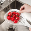 A hand is pouring water into a white kitchen strainer spoon filled with red tomatoes in a stainless steel sink.