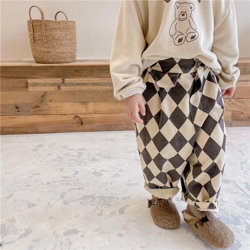 A child wearing beige long-sleeved top and checkered brown pants with pockets standing in front of a wooden shelf.
