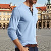 A man wearing a light blue long-sleeve casual shirt with a black collar and chest pocket, standing in front of historic Europ