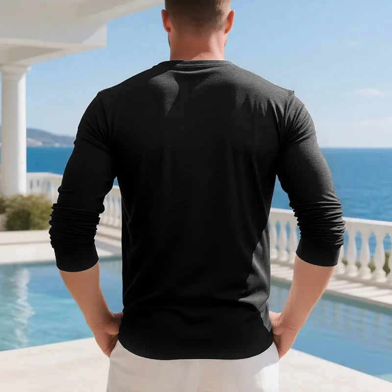 A man wearing a black long-sleeve shirt standing outdoors near a pool and ocean view.