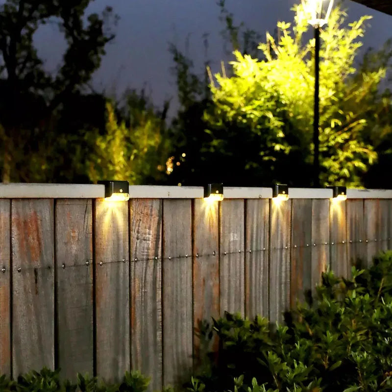 A row of solar powered deck lights illuminates a wooden fence and surrounding greenery at night.