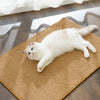 A white cat lying on a woven sisal scratching mat near a sunlit window.