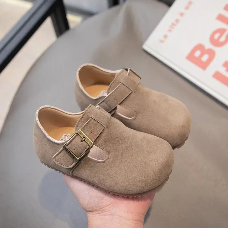 A pair of tan suede moccasin shoes with a leather hook-and-loop closure are held above a gray table.