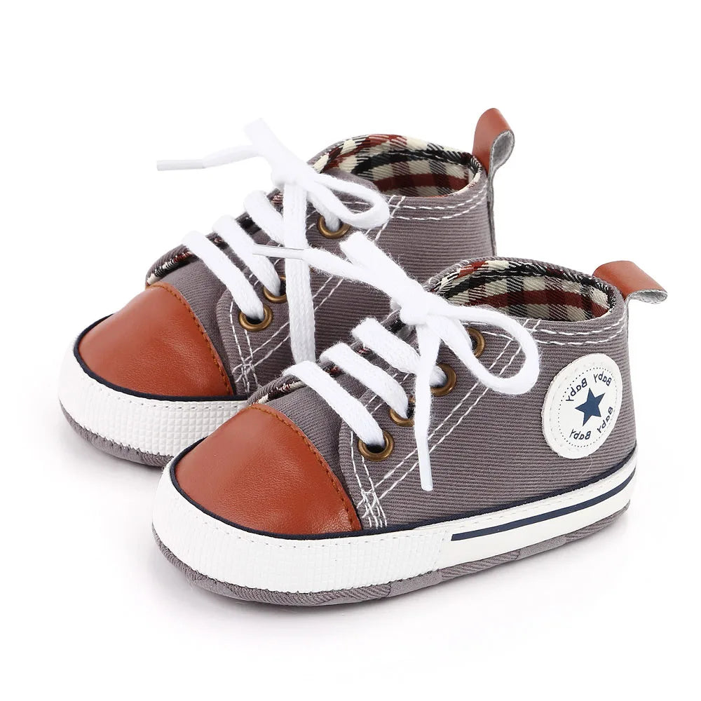 A pair of gray canvas baby sneakers with brown accents and white laces resting on a white background.