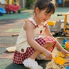 A young girl wearing a cream summer pinafore dress with a strawberry print and plaid skirt sitting on the ground.