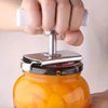 A hand is using a stainless steel manual tool to open a glass jar filled with orange-colored liquid.