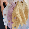 A yellow knitted cardigan sweater with a quilted pattern and pom-pom trim displayed on a wooden hanger.