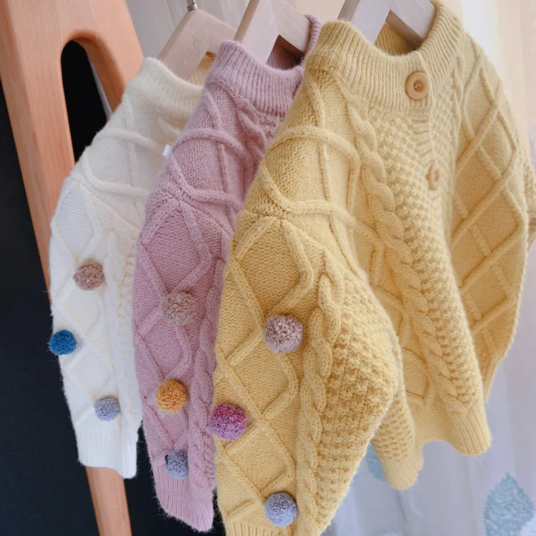 A yellow knitted cardigan sweater with a quilted pattern and pom-pom trim displayed on a wooden hanger.