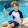 A smiling boy wearing a navy blue and white swimming set including a top, shorts, and cap, sitting by a pool.