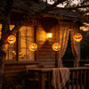 String lights with carved pumpkin lanterns hanging in front of a wooden house at dusk.