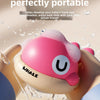 A pink and white whale-shaped baby bath toy is being used to spray water on a baby's hand during bath time.