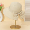 A beige straw sun hat with a large bow is displayed on a gold stand against a light background.