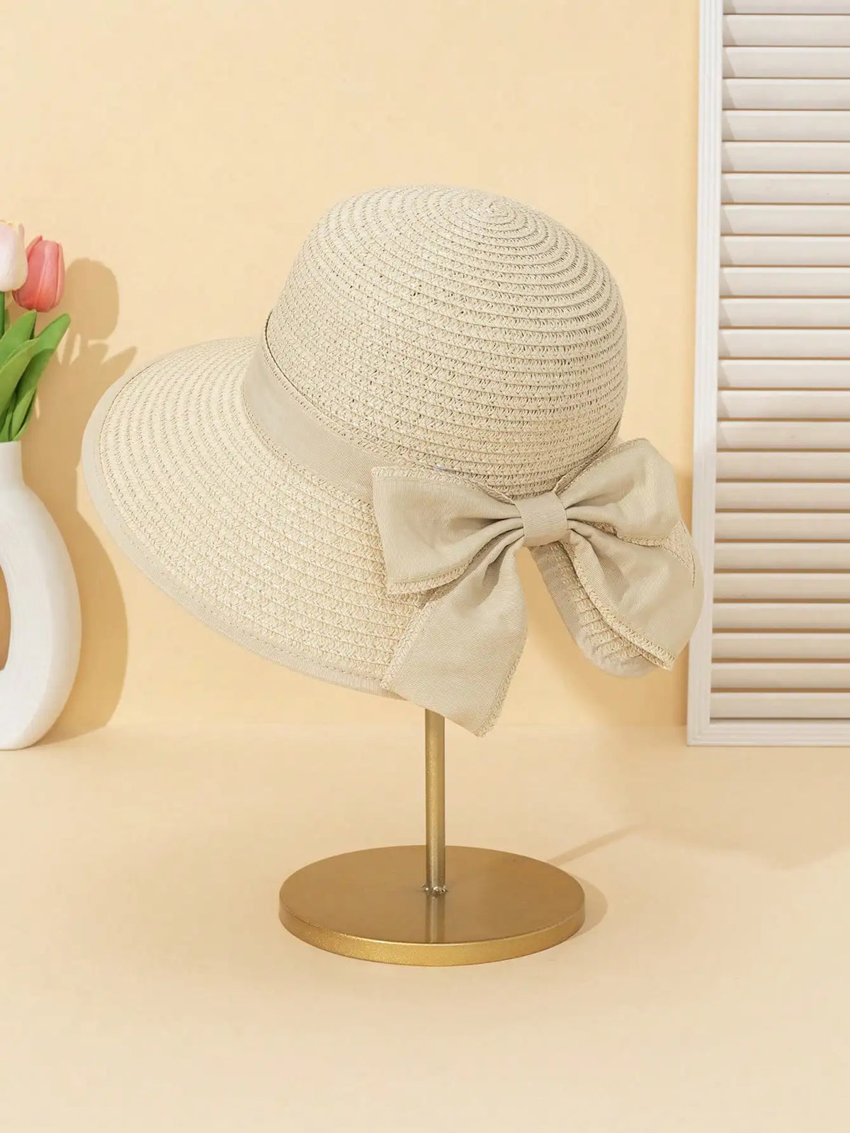 A beige straw sun hat with a large bow is displayed on a gold stand against a light background.