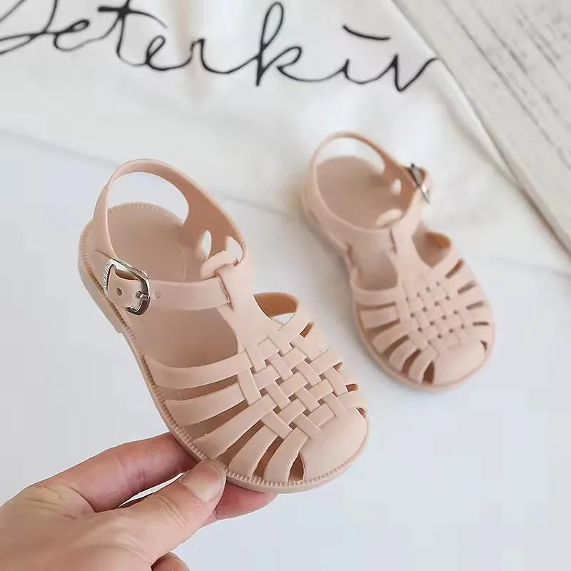 A pair of light pink kids' Roman Gladiator jelly sandals with woven straps and a non-slip sole being held by a hand.
