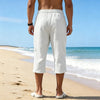 A man wearing white linen drawstring capri pants stands on a beach near the ocean.