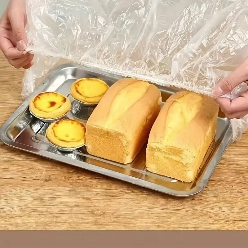 A clear plastic food cover is being placed over several yellow baked goods in a metal tray.