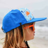 A close-up of a person wearing a blue baseball cap with an orange and blue print, sunglasses, and long blonde hair outdoors.