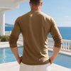 A man wearing a tan long-sleeve shirt and white pants stands by a pool with a blue ocean view.