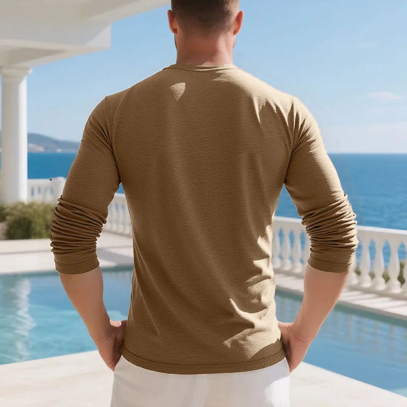 A man wearing a tan long-sleeve shirt and white pants stands by a pool with a blue ocean view.