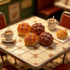 En tallerken med miniatyrbakverk i resin, inkludert croissanter og en kopp kaffe på et lyst bord.