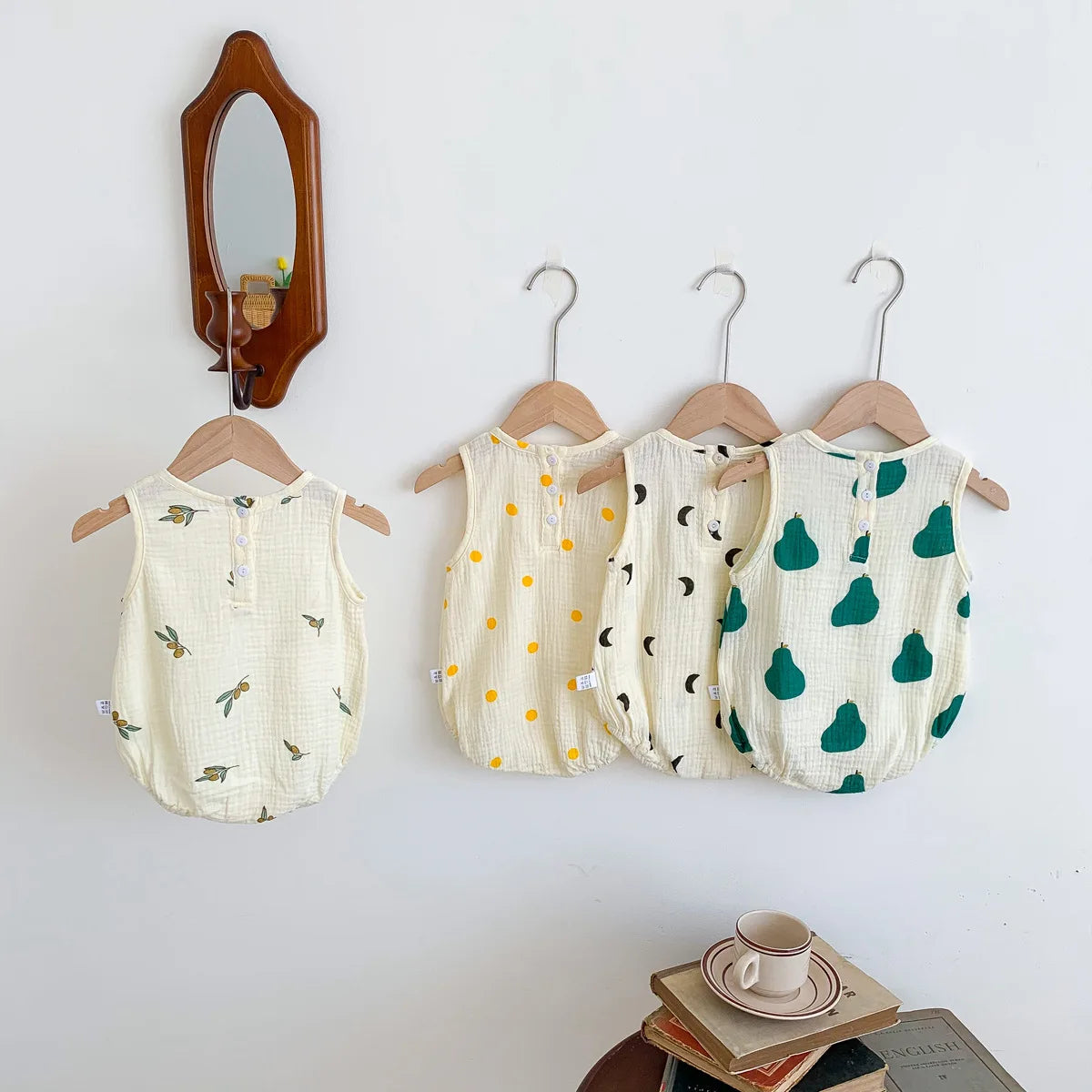 A set of baby cotton sleeveless jumpsuit bodysuits hanging on a wooden hanger against a white wall.