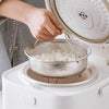 A hand places a stainless steel steamer basket into the inner pot of a white pressure cooker.