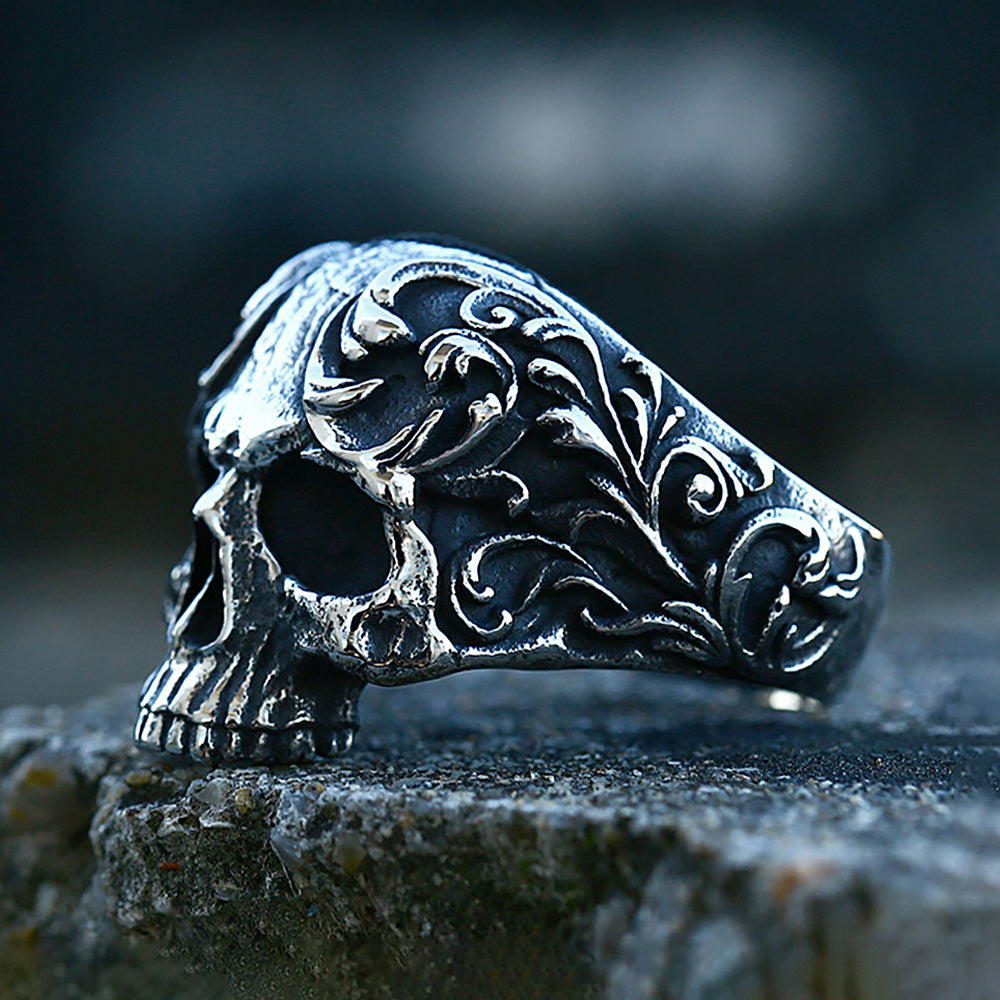 A silver gothic carved skull ring in 316L stainless steel resting on a dark stone surface.