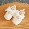 Pair of white lace baby first walker shoes with bow accents resting on a wooden surface.