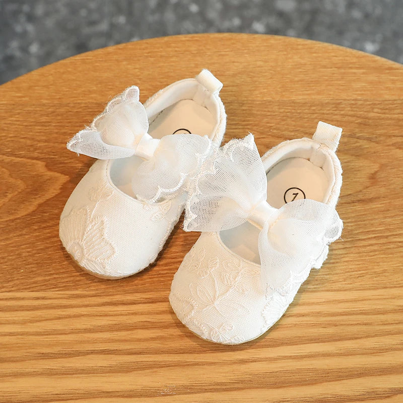 Pair of white lace baby first walker shoes with bow accents resting on a wooden surface.