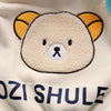 A close-up of a cream-colored fleece pajama top featuring a cute bear face with yellow ears and blue text below.