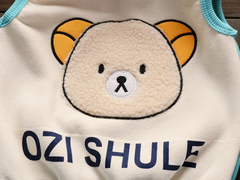 A close-up of a cream-colored fleece pajama top featuring a cute bear face with yellow ears and blue text below.