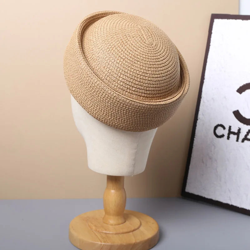 A beige straw pillbox sun hat displayed on a wooden mannequin against a light background.