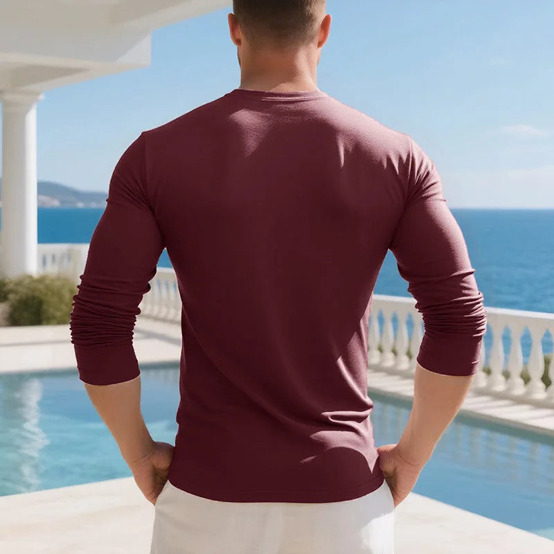A man wearing a maroon long-sleeve shirt standing outdoors by a pool and ocean view.