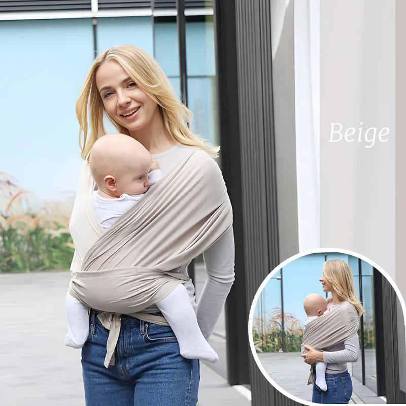 A blonde woman is wearing a beige baby wrap sling carrier while standing outdoors.