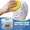 A hand holds a yellow cleaning cream sponge next to a white shoe cleaner jar and a white sneaker.
