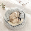 Lit cave moelleux gris pour animal avec doublure douce, posé sur une couverture blanche près d'une petite plante succulente.