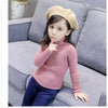 A young girl wearing a pink knit turtleneck sweater and dark pants sitting on a gray sofa.