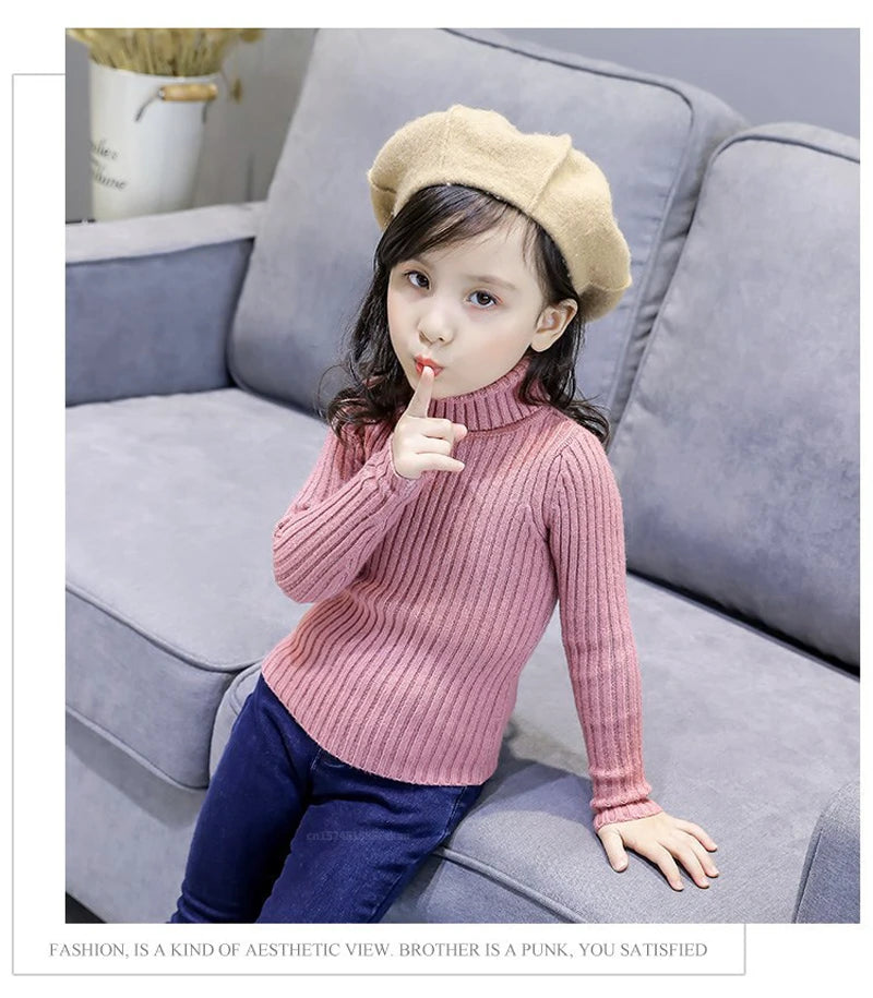 A young girl wearing a pink knit turtleneck sweater and dark pants sitting on a gray sofa.