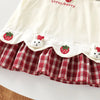 A white pinafore dress for kids featuring a strawberry and bunny print on the bodice and a red plaid skirt.