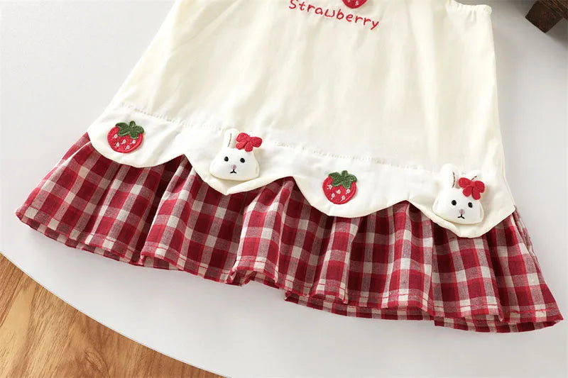 A white pinafore dress for kids featuring a strawberry and bunny print on the bodice and a red plaid skirt.