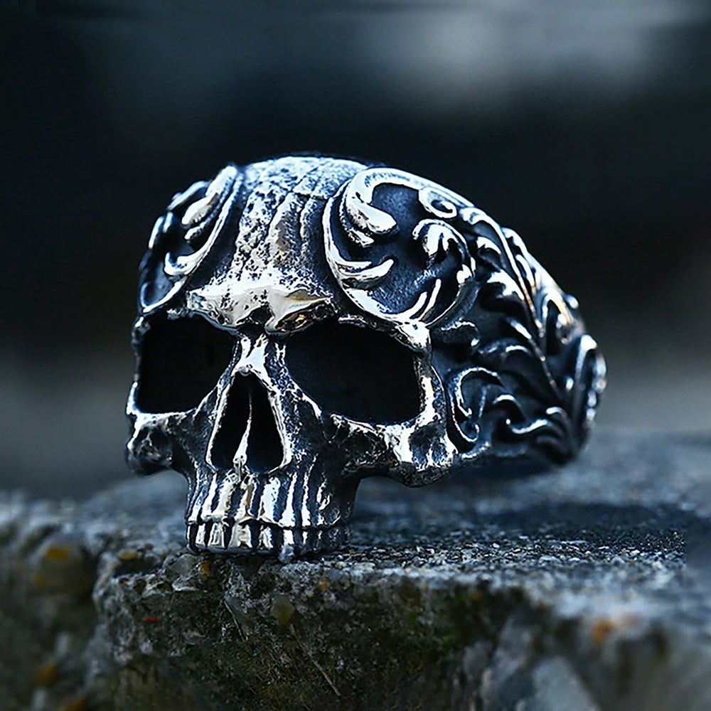 A dark, intricately carved gothic skull ring in stainless steel resting on a rough stone surface.