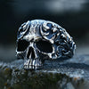 A dark, intricately carved gothic skull ring in stainless steel resting on a rough stone surface.