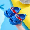 A pair of blue kids shark clog sandals with red teeth on the toe, shown against a bright yellow background.